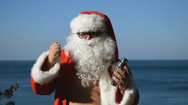 Bir adam Noel Baba kostümlü kokteyl shaker ile sahil üzerinde dans ediyor. Seyahat ve tatil