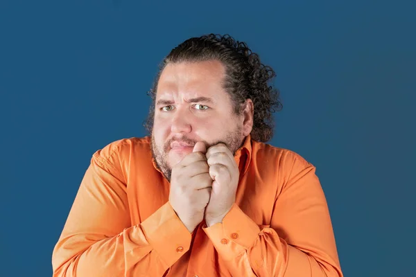 Grubas w shirt pomarańczowy posiada jego ręce nad jego twarz na niebieskim tle. On jest bardzo zaskoczony. — Zdjęcie stockowe
