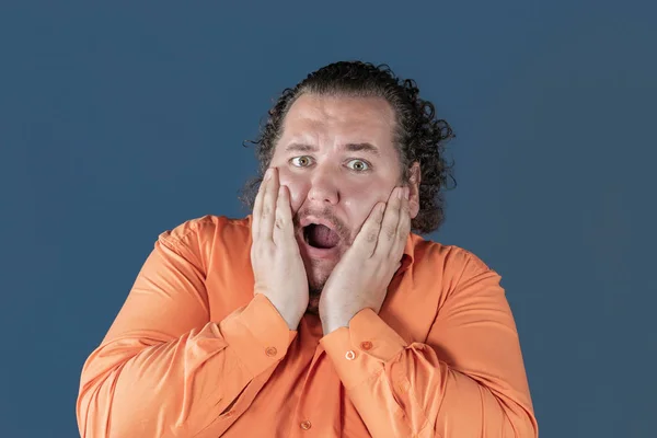 Hombre gordo con camisa naranja sostiene sus manos sobre su cara sobre fondo azul. Está muy asustado. — Foto de Stock