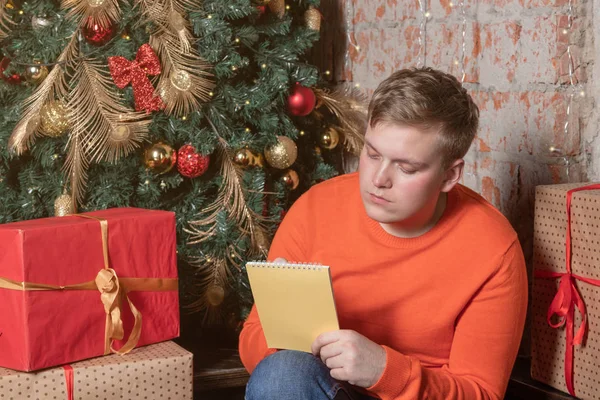 Handsome guy is writing a letter to Santa sitting under the tree surrounded by boxes of gifts