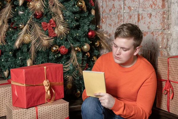Handsome guy is writing a letter to Santa sitting under the tree surrounded by boxes of gifts