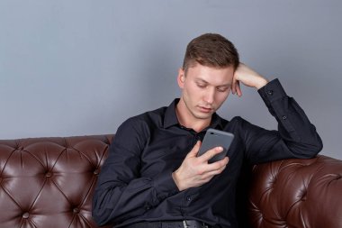Siyah gömlek giyen çekici genç yakışıklı adam akıllı telefon tutan deri kanepede oturan. Konfor ve rahatlama.