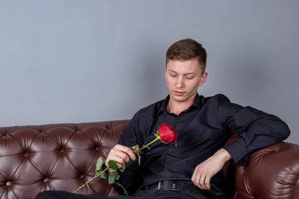 Hombre guapo con camisa negra sentado en el sofá de cuero sosteniendo una rosa y preparándose cita romántica —  Fotos de Stock