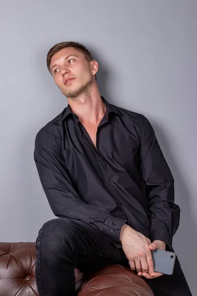 Atraktivní mladý pohledný muž na černé košili, který sedí na kožené pohovce a drží telefon. Pohodlí a relaxace. — Stock fotografie