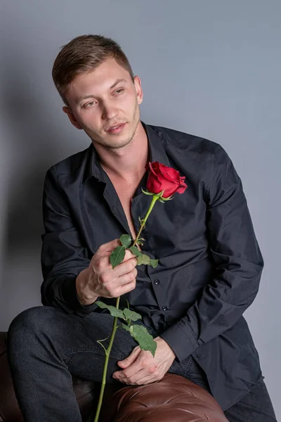 Stilig man bär svart skjorta sitter på skinnsoffan håller en ros och få redo romantiskt datum — Stockfoto