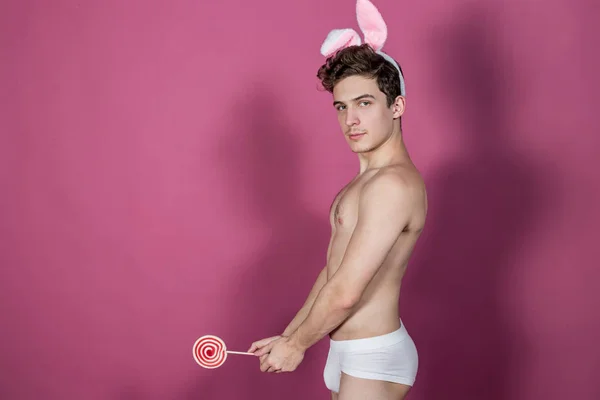 Смішний голий хлопець у вухах кролика тримає льодяник на рожевому фоні — стокове фото
