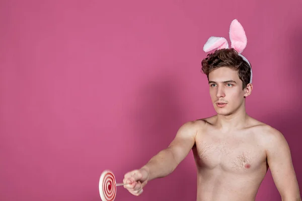 Veselý nahý chlápek v králíčcích, který drží lízáka přes růžové pozadí — Stock fotografie