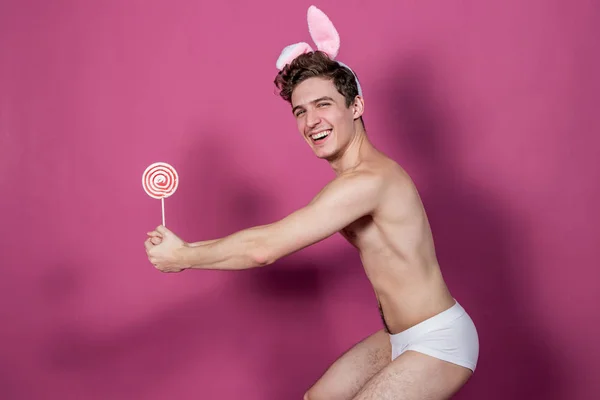 Смішний голий хлопець у вухах кролика тримає льодяник на рожевому фоні — стокове фото