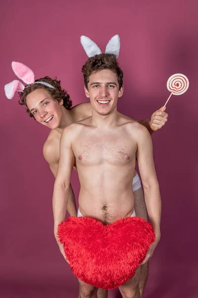 Двоє смішних голих хлопців у вухах кролика тримають льодяник і велике червоне серце над рожевим тлом — стокове фото