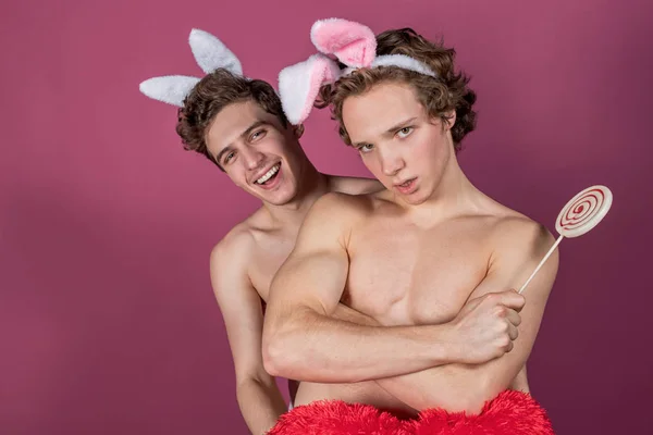 Dva vtipní nahatí chlápci s králíčkama držící lízáka a velké rudé srdce na růžovém pozadí — Stock fotografie