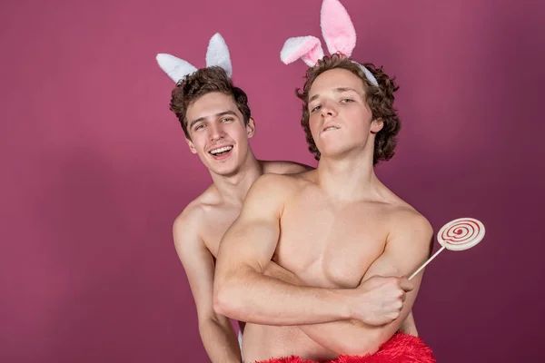 Двоє смішних голих хлопців у вухах кролика тримають льодяник і велике червоне серце над рожевим тлом — стокове фото