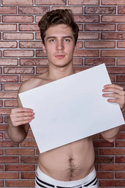 Young naked sexy guy posing in underwear with a billboard on the background of a brick wall