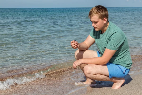 Hombre joven en una camiseta en la orilla del mar y mira al mar, vista trasera —  Fotos de Stock