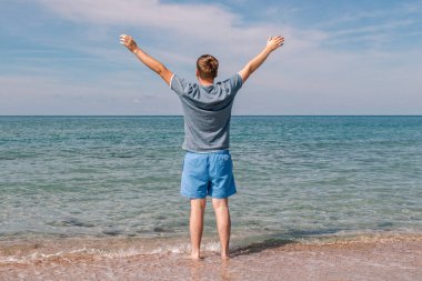 Young man in a t-shirt on the seashore and looks at the sea, back view clipart