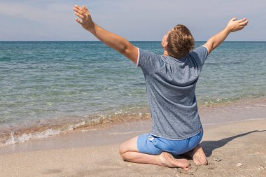 Sahilde oturan bir t-shirt mutlu genç adam ve denize bakıyor, arka görünüm