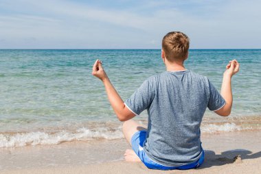 Plajda bacaklarla oturan mutlu genç adam. Yoga ve Fitness