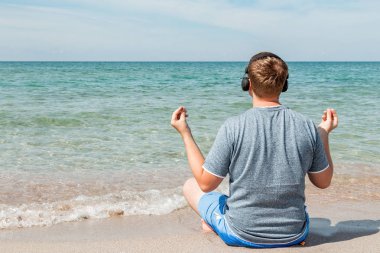 Plajda oturan mutlu genç adam kulaklıklar müzik dinlerken. Yoga ve dinlenmek
