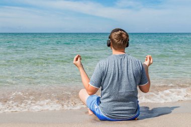 Plajda oturan mutlu genç adam kulaklıklar müzik dinlerken. Yoga ve dinlenmek