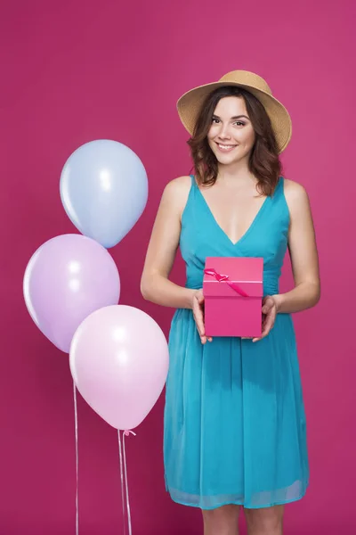 Chica en un sombrero de paja y vestido azul sostiene un regalo en sus manos sobre un fondo rosa — Foto de Stock