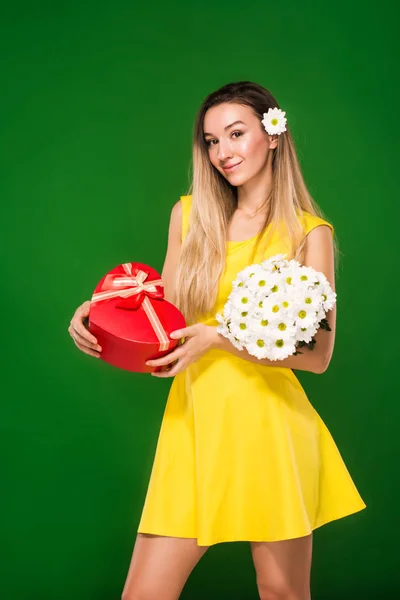 Chica rubia en un vestido amarillo sostiene un ramo de manzanillas y una caja de regalo — Foto de Stock