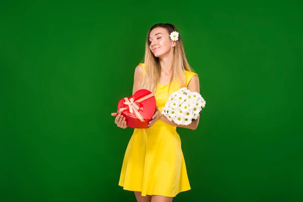 Chica rubia en un vestido amarillo sostiene un ramo de manzanillas y una caja de regalo — Foto de Stock
