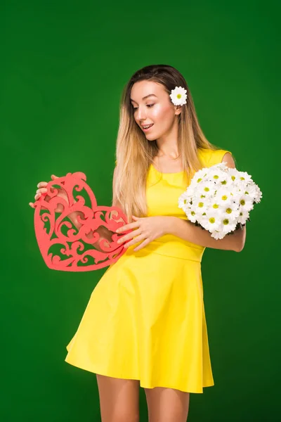 Hermosa chica rubia en un vestido amarillo sostiene un corazón en sus manos y un ramo de manzanillas — Foto de Stock
