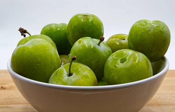 Close up of green plums fruit in a ceramic jar. Wooden table, white background