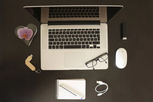Office objects composition, black desk, laptop, mouse, USB flash drive, portable recharge, sketch pad, pen, cable, eyeglasses, purple flower-shaped candle, top view