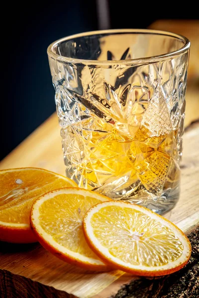 Closeup view on rum or whiskey in a glass with ice on a wooden background with sliced orange. Alcohol glass bar. Copy space for brand, text or logo.