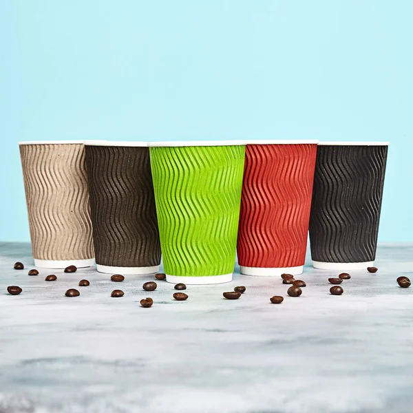 multicolored coffee cup with coffee beans on blue background. disposable paper dishes. Brown, red, green, black cups. Copy space for logo, text or brand
