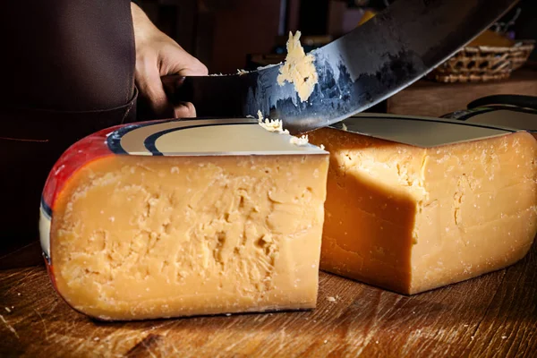 Young worker man cuts half of wheel gauda cheese in shop. Wooden background, knife for cheese. Close up view. Copy space for text, logo or brand. Work Concept.