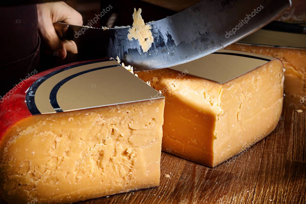 Young worker man cuts half of wheel gauda cheese in shop. Wooden background, knife for cheese. Close up view. Copy space for text, logo or brand. Work Concept.