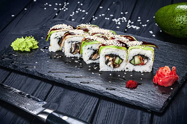 Sushi rolls with avocado, unagi sauce and eel on dark stone served with ginger and wasabi and wooden black wooden table. Fresh Japanese cuisine. Close up view on asian food. Sushi image for menu.