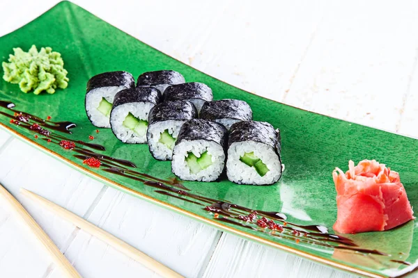 Maki rolls with cucumber on green plate served with ginger and wasabi on white wooden table. Fresh Japanese cuisine. Close up view on asian food. Sushi image for menu.