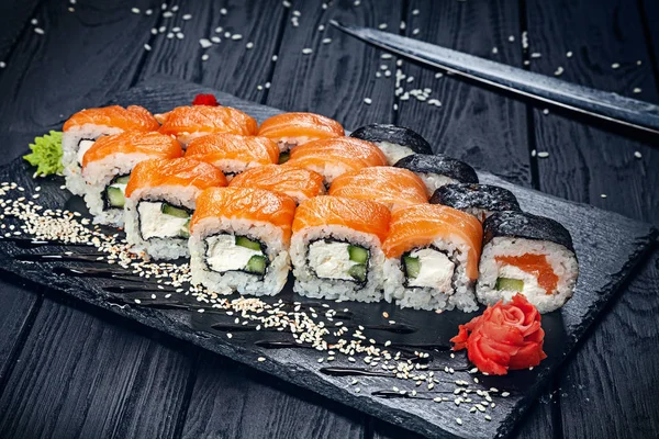 A set of sushi rolls with salmon on dark stone served with ginger and wasabi and wooden black wooden table. Fresh Japanese cuisine. Close up view on asian food. Sushi image for menu.