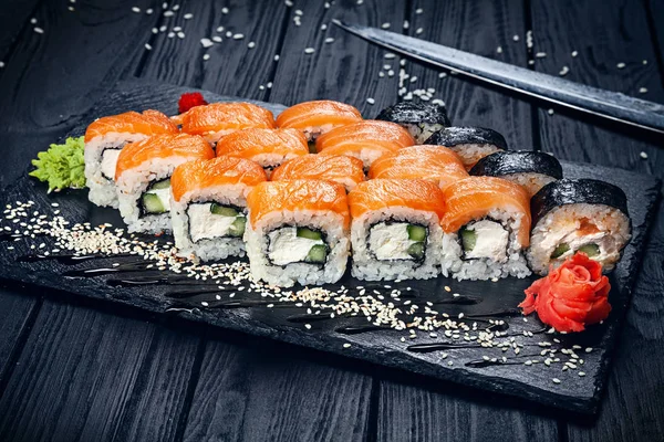 A set of sushi rolls with salmon on dark stone served with ginger and wasabi and wooden black wooden table. Fresh Japanese cuisine. Close up view on asian food. Sushi image for menu.