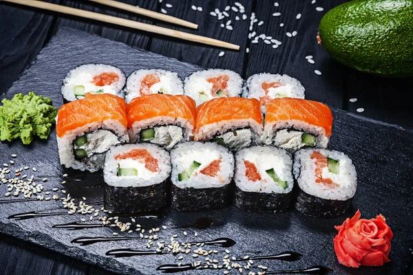 A set of sushi rolls with salmon on dark stone served with ginger and wasabi and wooden black wooden table. Fresh Japanese cuisine. Close up view on asian food. Sushi image for menu.