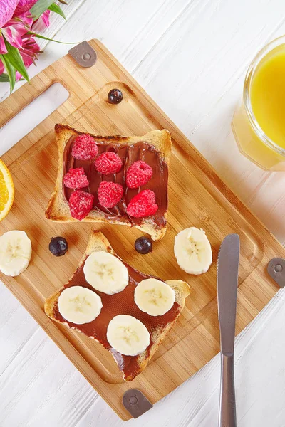 Top view on toast with Peanut butter and raspberries, banana. Breakfast Composition flat lay of orang juice, flowers and toast on white wooden background. Traditional summer breakfast. copy space — Stock Photo, Image