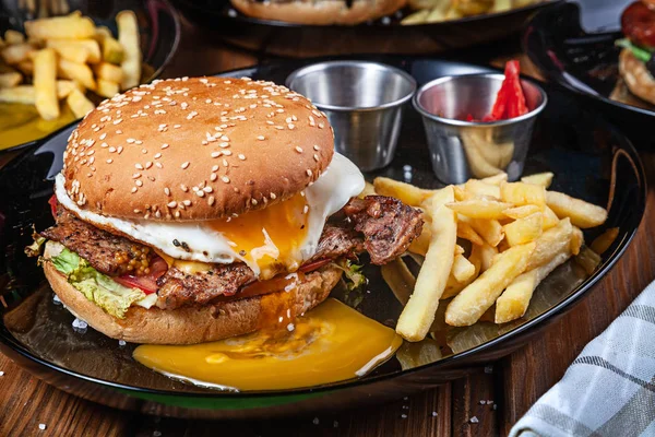 Saftige und leckere Burger mit Ei, Salat und Soße auf einem schwarzen Teller mit Pommes frites. Amerikanisches Fast Food. Hamburger mit Kopierraum. Nahaufnahme, selektiver Fokus. Sommerkost. Grillmenü — Stockfoto