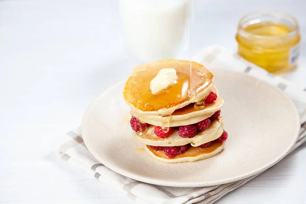 Nahaufnahme auf einem Stapel frischer, hausgemachter Pfannkuchen mit Himbeeren, schmelzender Butter und dem Gießen von Honig oder Marmorsirup. Amerikanischer Pfannkuchen auf weißem Hintergrund mit Morgenlicht. Frühstück. Selektiver Fokus. — Stockfoto