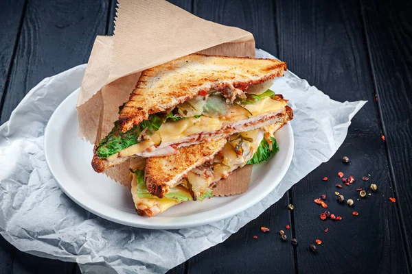 Close up on grilled sandwich with chicken and melted cheese and lettuce. Snack. Fast food for lunch. Sandwich served on white plate on dark wooden background