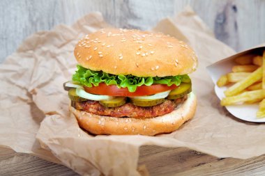 Hamburgerin yakın görüntüsü, kahverengi kağıtta patates kızartması. Geleneksel fast food. Salatalı, domatesli, peynirli ve salatalıklı taze lezzetli hamburger. Tasarım için alanı kopyala. Sağlıksız yiyecekler. Şişko