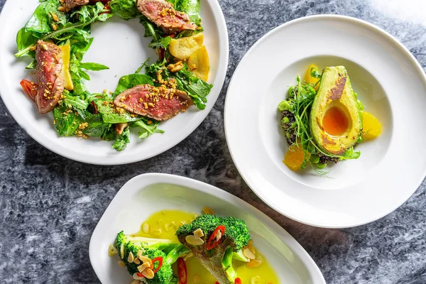 Top view green style salad on marble table. Three different salad with beef tataki, grilled avocado and brocolli on white plate. Tasty restaurant food style. Flat lay for recipe or banner