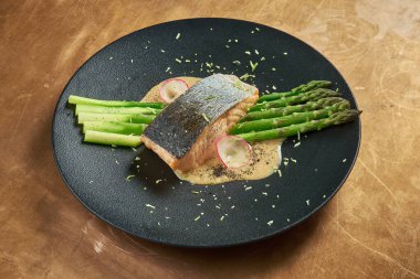 Fırında somon bifteği, ızgara kuşkonmaz ve siyah bir tabakta beyaz sos, bakır bir arka planda. Restoran servisi.