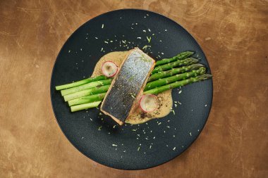 Fırında somon bifteği, ızgara kuşkonmaz ve siyah bir tabakta beyaz sos, bakır bir arka planda. Restoran servisi.