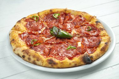 Ev yapımı pizza, sucuklu salam, acı biber ve erimiş peynir beyaz bir tabakta beyaz ahşap arka planda.