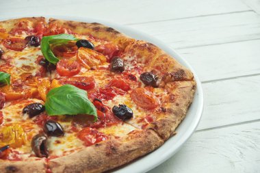 İştah açıcı, ev yapımı margaritalı pizza kiraz domatesli, fesleğenli ve mozzarellalı beyaz bir tabakta ahşap arka planda.