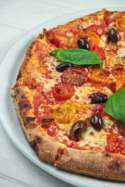 İştah açıcı, ev yapımı margaritalı pizza kiraz domatesli, fesleğenli ve mozzarellalı beyaz bir tabakta ahşap arka planda.