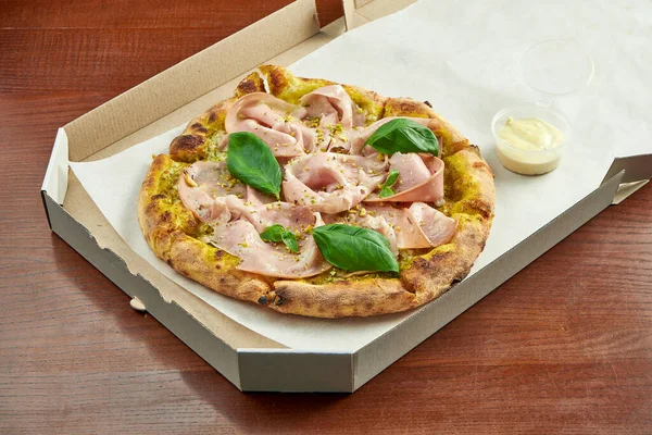 Photo for pizza delivery. Pizza with mortadella sausage, pistachios, basil and melted cheese in a cardboard box with side sauce on a wooden table