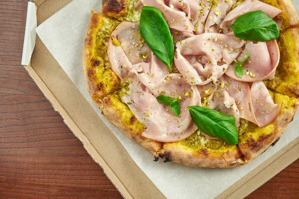 Photo for pizza delivery. Pizza with mortadella sausage, pistachios, basil and melted cheese in a cardboard box with side sauce on a wooden table